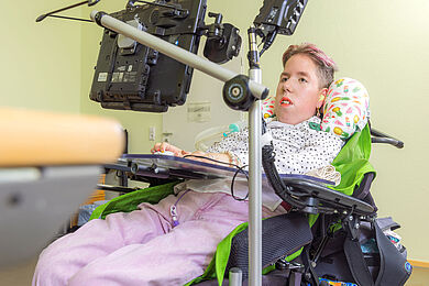 Eine Teilnehmerin sitzt im Rollstuhl und schaut auf einen Bildschirm. Der Rollstuhl sieht sehr modern aus und bietet viele technische Hebel und Knöpfe. Die Teilnehmerin trägt eine rosa Jeans, eine weiße Bluse mit schwarzen Punkten und hat pinke Strähnen in den Haaren, die sie kurz trägt. Sie ist uns zugewandt. Im Hintergrund erkennt man eine hellgrüne Wand.