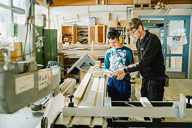 2 Männer stehen in einer Holzwerkstatt. Der eine Mann erklärt dem anderen Mann etwas. 