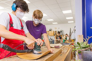 Mitarbeiter und Werkstattmitarbeiter schleifen eine Stuhllehne aus Holz ab. Beide tragen Brillen und Masken.