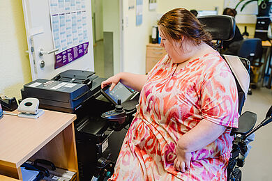 Eine Teilnehmerin in einem weiß-roten Kleid im Rollstuhl bedient einen großen Drucker. 