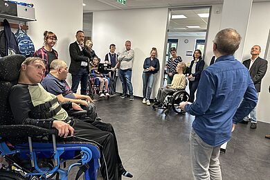 Ein Mann mit blauem Hemd ist in Rückenansicht zu sehen. Er scheint zu den anderen Personen zu sprechen, die im Halbkreis um ihn herumstehen und sitzen. Es sind 4 Personen im Rollstuhl zu sehen.