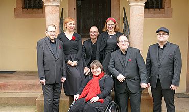 Mitglieder des inklusiven A-Capella-Ensembles Thonkunst vor der St. Gangolf-Kirche in Kohren-Salis. Alle tragen schwarze Kleidung mit roten Accessoires. 