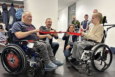 Ein rotes Band wird von zwei Rollstuhlfahrern gehalten. Dazwischen hocken ein Mann und eine Frau vor dem Band. Der Mann schneidet das rote Band durch.