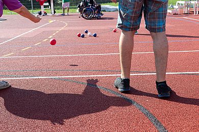 Auf dem Bild sind Menschen beim Boule-Spiel zu sehen. Im Hintergrund stehen zwei Rollstühle.