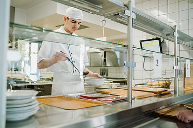 Das Bild zeigt einen jungen Mann in einer Großküche an der Essensausgabe. Er trägt weiße Kochkleidung. Mit der einen Hand schöpft er Suppe in eine Schale, die er in der anderen Hand hält. Er sieht sehr konzentriert aus. 
