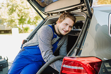 Ein Teilnehmer sitzt in einem geöffneten Kofferraum und schaut mit einem leichten Lächeln in die Kamera. In der Hand erkennt man einen Schlauch eines Staubsaugers. Er trägt eine blaue Arbeitshose und darunter ein graumeliertes, langärmeliges Shirt.