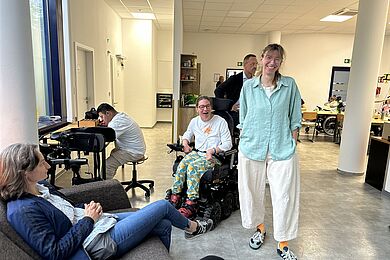 Person sitzt links unten auf einem Sofa. Mittig eine Frau mit weißer Hoseund hellblauer Bluse, die in die Kamera lacht. Links davon eine Frauim Rollstuhl, die fröhlich aussieht. Im Hintergrund weitere Personen.