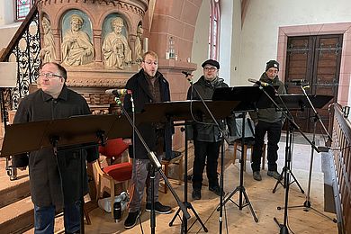 Mitglieder des inklusiven A-Capella-Ensembles Thonkunst tragen warme Kleidung und singen in einer Kirche. Vor ihnen sind Mikrofone und Notenständer aufgebaut.