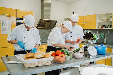 3 Personen arbeiten in einer Küche mit gelben Schrankfronten. Sie stehen nebeneinander an einem mittig platzierten Arbeitstisch und sind auf ihrer jeweiligen Tätigkeiten konzentriert. Sie tragen weiße Kochuniformen. Im Vordergrund steht ein Korb mit Brötchen, eine Schüssel mit Gemüse und eine Waage.