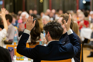 Ein Gast mit einem dunklen Jackett über einem weißen Hemd von hinten. Er applaudiert in Gebärdensprache, in dem er beide Hände neben dem Kopf erhoben bewegt. Im Hintergrund erkennt man unscharf weitere Gäste, die das auch tun.