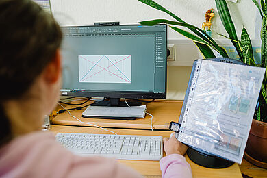 Das Bild ist über die Schulter einer Teilnehmenden aufgenommen. Sie sitzt an einem Schreibtisch und schaut auf einen Bildschirm. Darauf ist ein Bildbearbeitungsprogramm zu erkennen, sie arbeitet an einer Vorlage mit verschiedenfarbigen Linien. Rechts neben ihr ist ein Aufsteller zu sehen, in dem ein Dokument in einer Klarsichtfolie ist. 