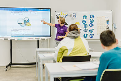 Eine Mitarbeiterin in einem lila T-Shirt mit kinnlangen hellbraunen Haaren deutet mit der rechten Hand auf ein Bild auf einem großen Bildschirm. Vor ihr sitzen an Tischen zwei Teilnehmende. Wir sehen die Teilnehmenden von hinten und die Frau von vorne. Auf dem Bildschirm sieht man einen Helm, Kopfhörer und Handschuhe. Neben der lehrenden Person hängt rechts ein Plakat mit verschiedenen Mitteln zur Arbeitssicherheit.