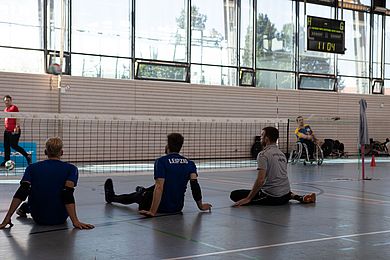 In einer Turnhalle sitzen drei Männer vor einem Volleyballnetz. Ein wenig abseits steht ein Rollstuhlfahrer.