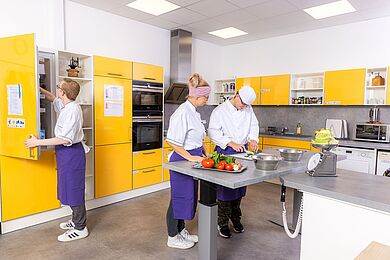 Drei Personen in einer Küche mit gelben Fronten. Alle tragen weiß-blaue Kochkleidung. Ein Mitarbeiter schneide eine Gurke, eine Frau schaut ihm zu. Ein anderer Mitarbeiter steht am geöffneten Kühlschrank.