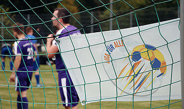 In einem Fußballtor hängt ein kleines Banner mit dem Logo und dem Schriftzug "Liga für alle". Im Hintergrund sieht man verschwommen Fußballspieler*innen.