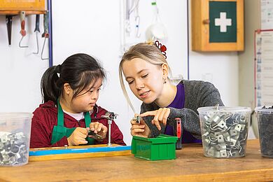 Eine Mitarbeiterin mit blonden Haaren, die sie mit einer Spange hochgesteckt hat, erklärt einer Werkstattmitarbeiterin ein Metallteil