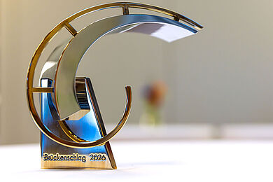 Brückenschlagpreis 2026: Eine vergoldete Skulptur, die optisch einer Brücke nachempfunden ist. Auf dem Sockel steht "Brückenschlag 2026".